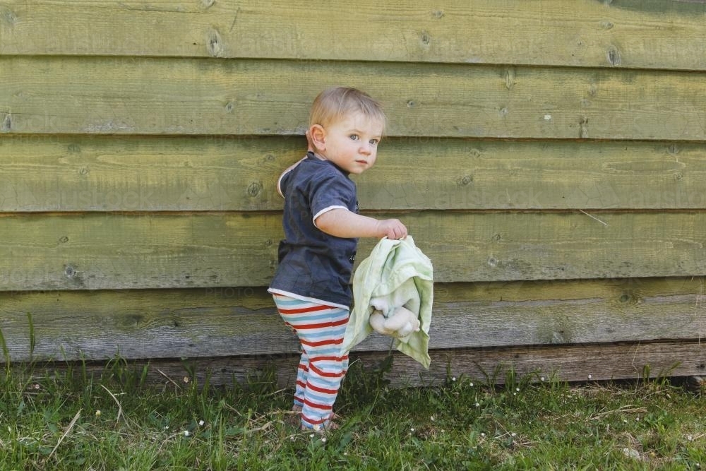 Toddler boy outside with his teddy beside green slat wall - Australian Stock Image