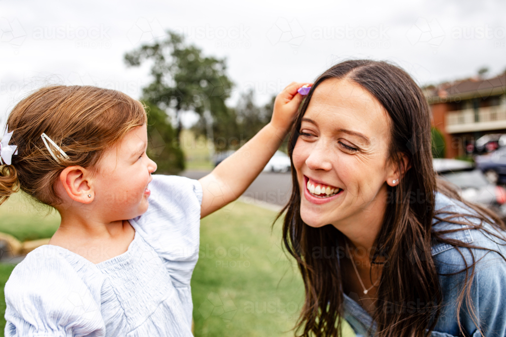 Toddler daughter putting tiny purple flower on mothers hair. - Australian Stock Image