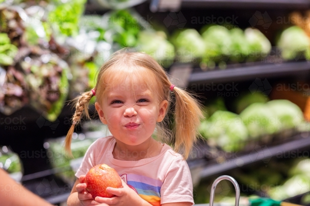 Toddler girl eating fresh red apple in grocery store with cheeky look - Australian Stock Image