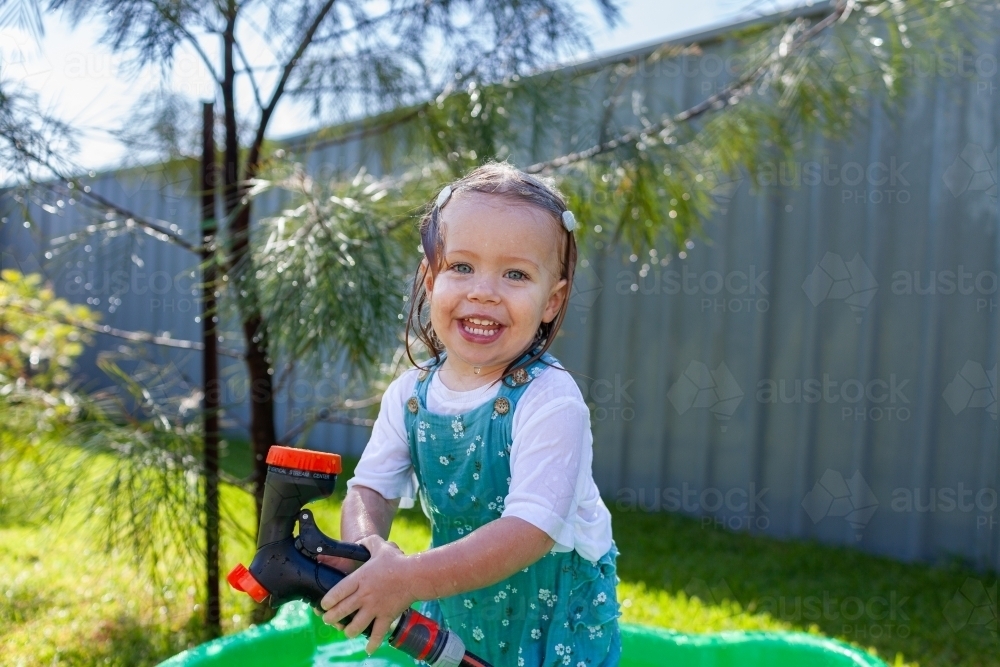 Toddler girl in suburban backyard playing with hose and getting wet in summertime - Australian Stock Image