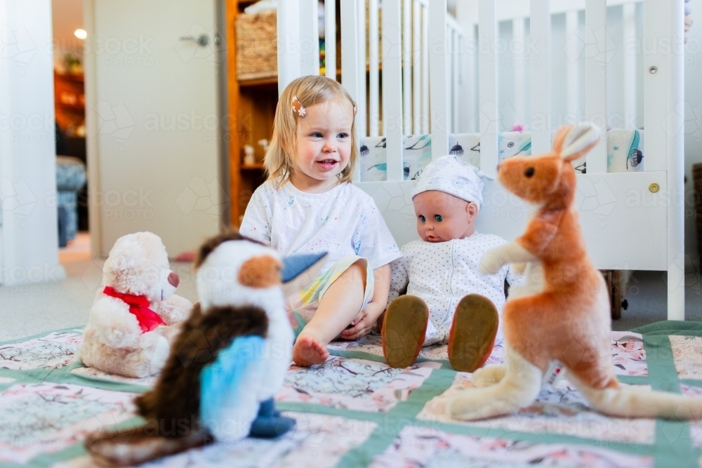 Toddler girl playing picnic with dolly and soft toys on mat in bedroom - Australian Stock Image