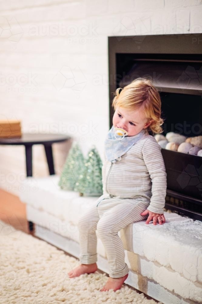 Toddler in pyjamas sitting inside, with a dummy in his mouth - Australian Stock Image