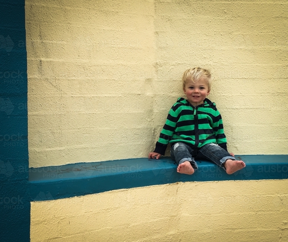 Toddler in stripey jumper sitting against yellow wall smiling at camera - Australian Stock Image