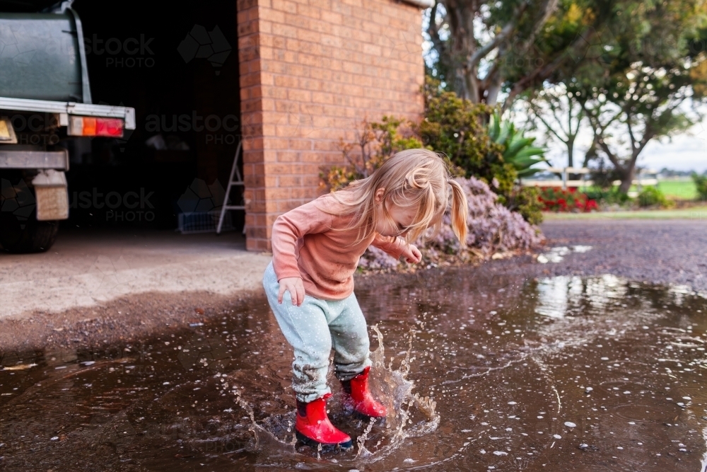Toddler jumping in rain water puddle on farm in red gumboots - Australian Stock Image