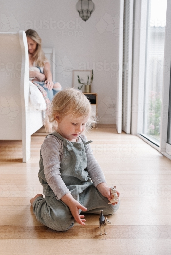 Toddler playing on the floor while mother feeds her sibling. - Australian Stock Image