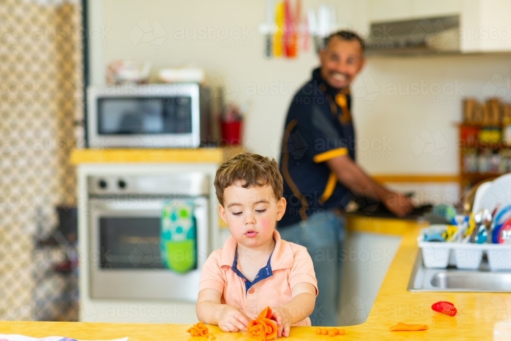 toddler playing with play dough while father looks on from kitchen - Australian Stock Image