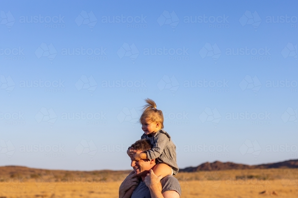 toddler riding on father's shoulders against blue Pilbara sky - Australian Stock Image