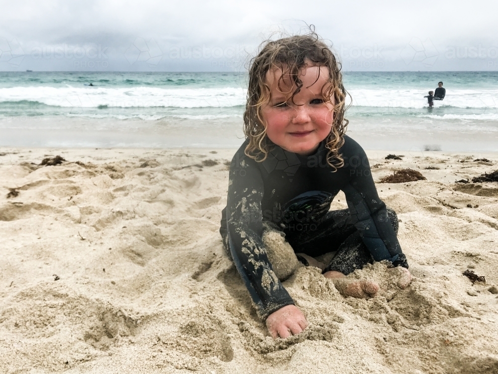 Toddler sitting on beach in wetsuit playing in sand looking at camera smiling - Australian Stock Image