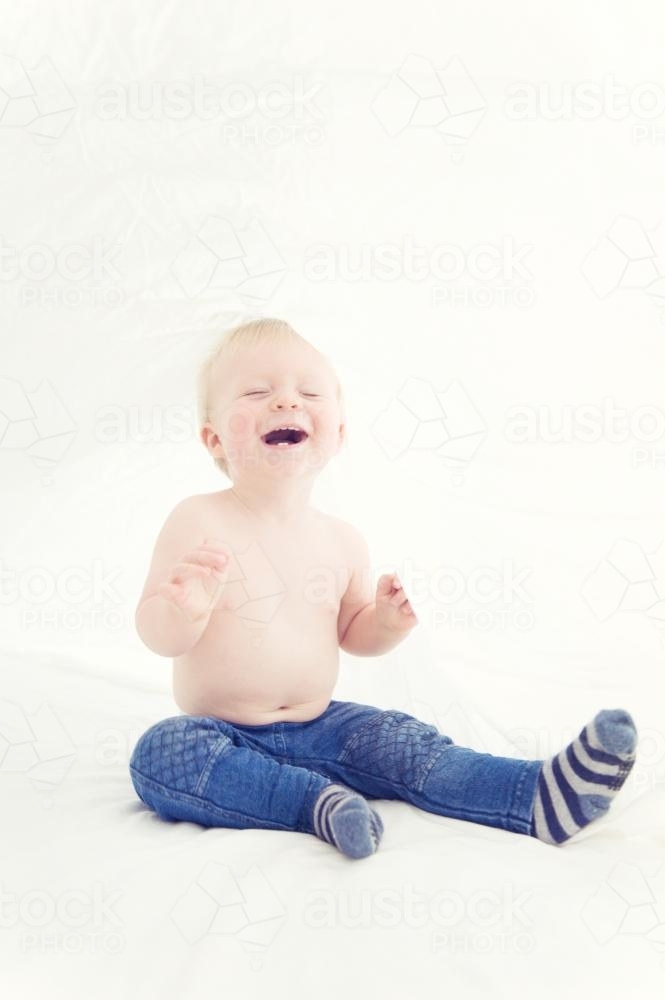 Toddler sitting on bed under the sheets wearing jeans and socks laughing - Australian Stock Image