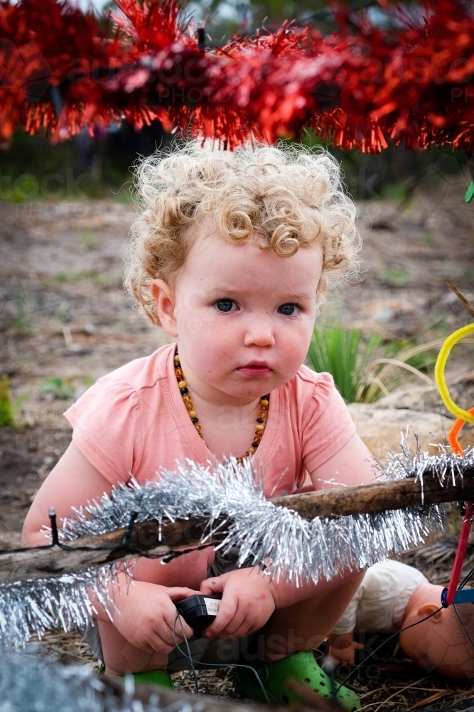 Toddler squatting down behind Bush Christmas tree decorated in tinsel - Australian Stock Image