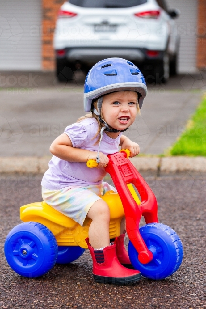 Toddler with cheeky grin riding tricycle on quiet road - Australian Stock Image