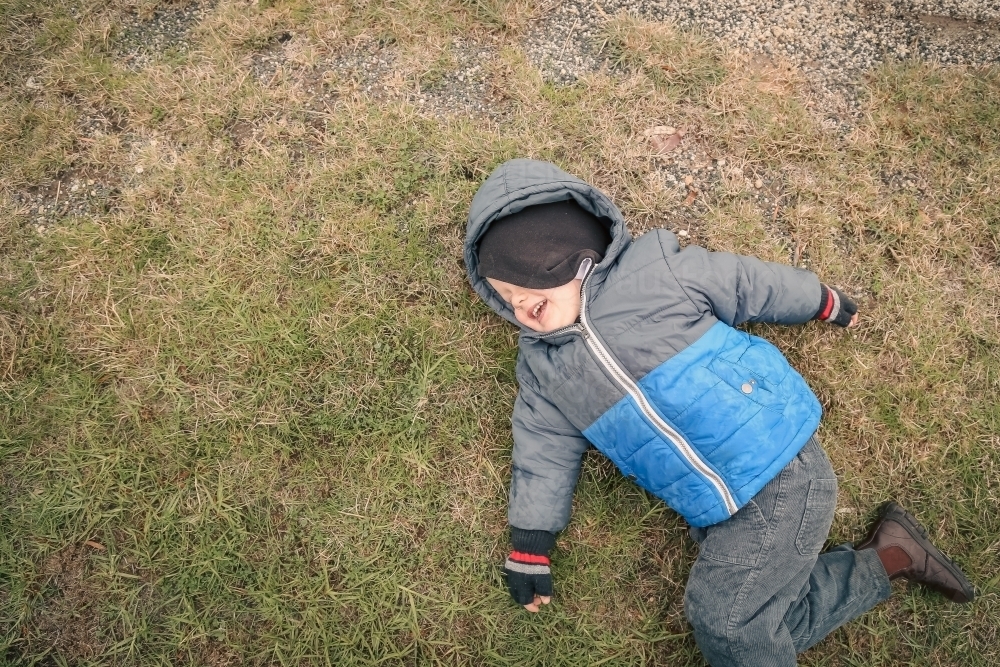 Top down view of little boy lying on grass in winter with cheeky smile - Australian Stock Image