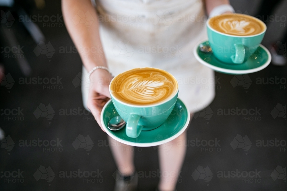 Top view shot of 2 cups of coffee with an art in a green cup being carried by a woman - Australian Stock Image