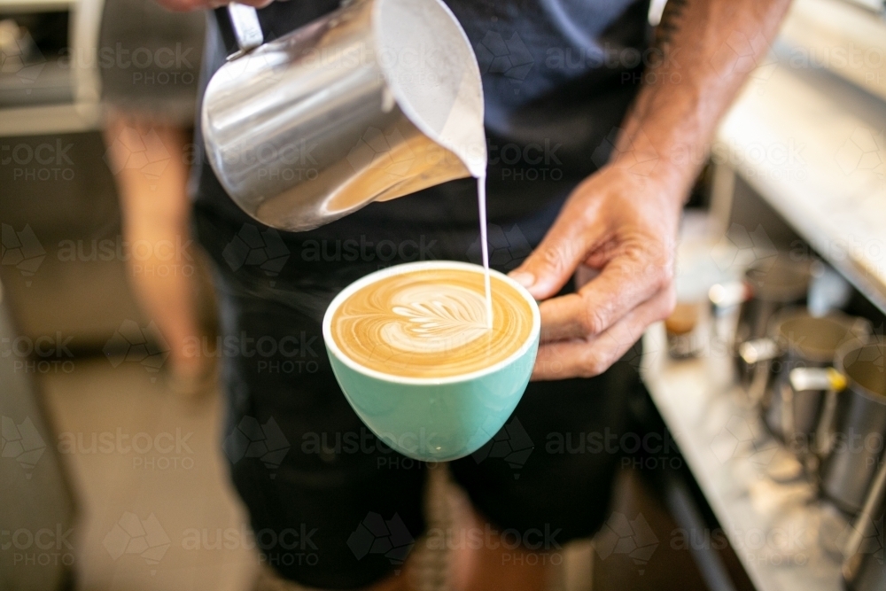 Top view shot of a barista pouring milk from a tin jar to the coffee in a green mug - Australian Stock Image