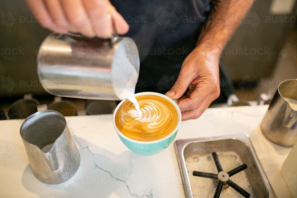Top view shot of a barista pouring milk from a tin jar to the coffee in a green mug making latte art - Australian Stock Image