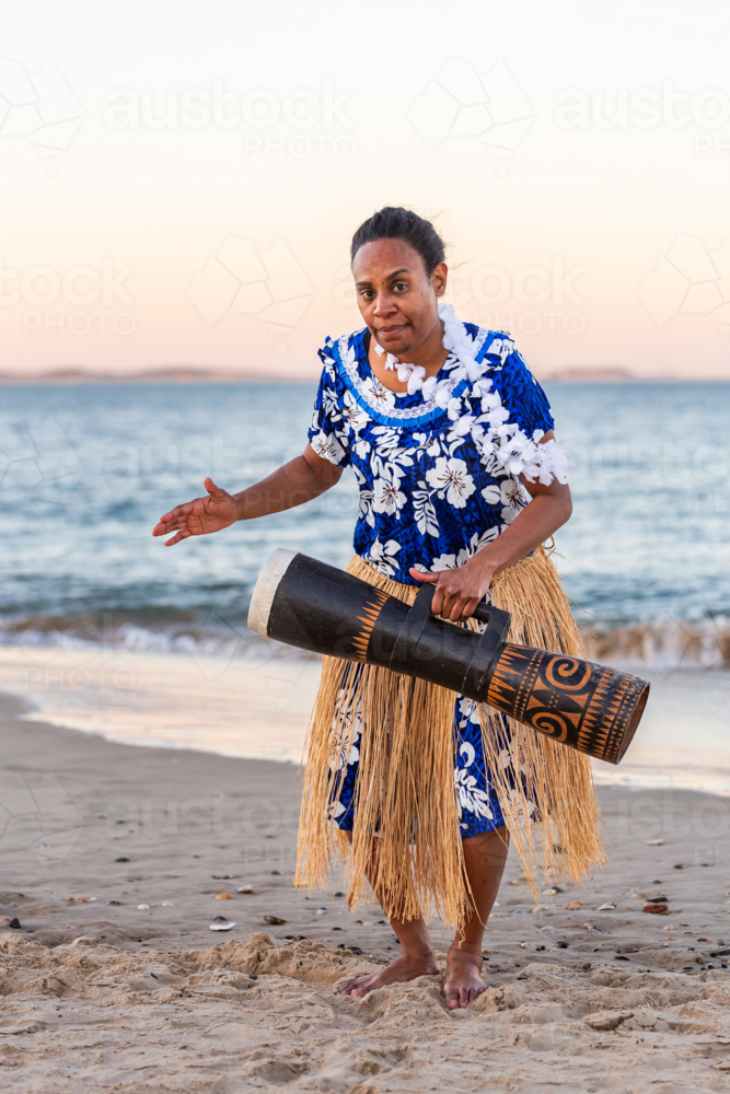 Torres Strait Islander dance performer in traditional floral dress holding drum by ocean - Australian Stock Image