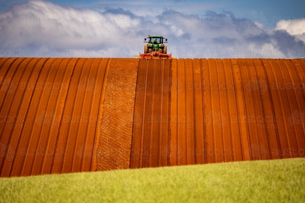 Tractor cultivating red soil in hilly paddock with green cereal crop in foreground, horizontal view - Australian Stock Image
