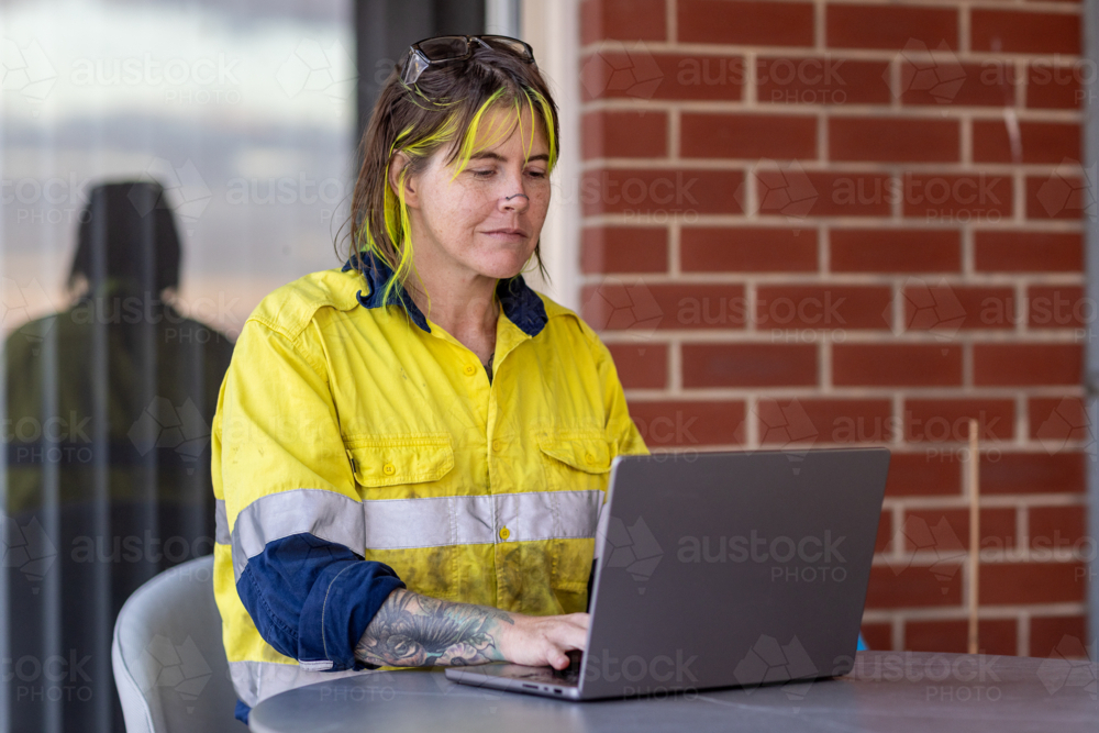 tradie wearing hi-vis shirt with reflective strips sitting at table working on laptop computer - Australian Stock Image