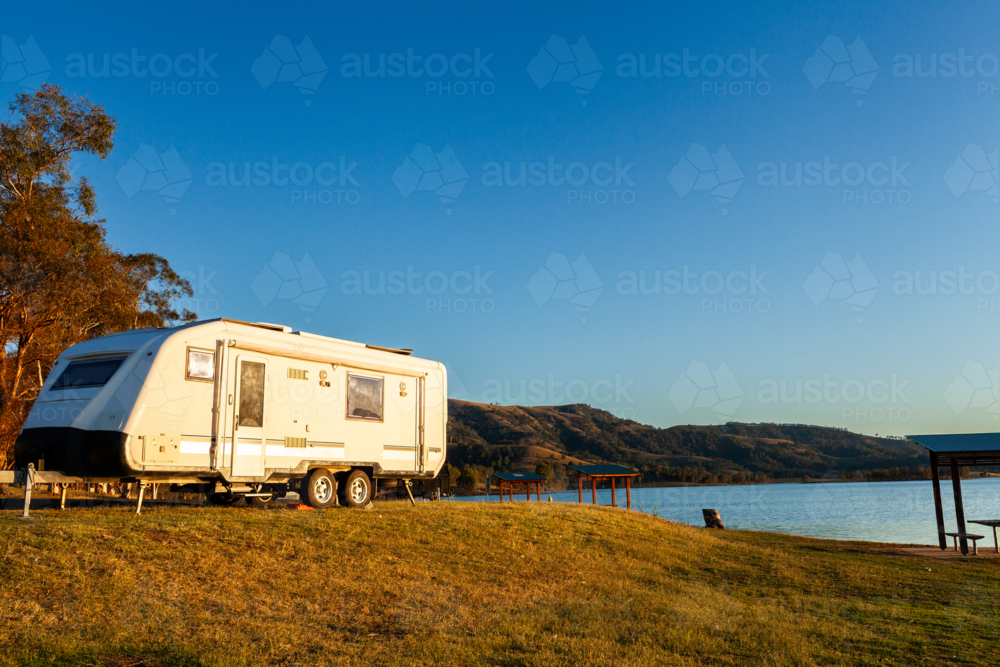 Travel concept – caravan parked by lakeside in sunset light with blue sky copy space - Australian Stock Image
