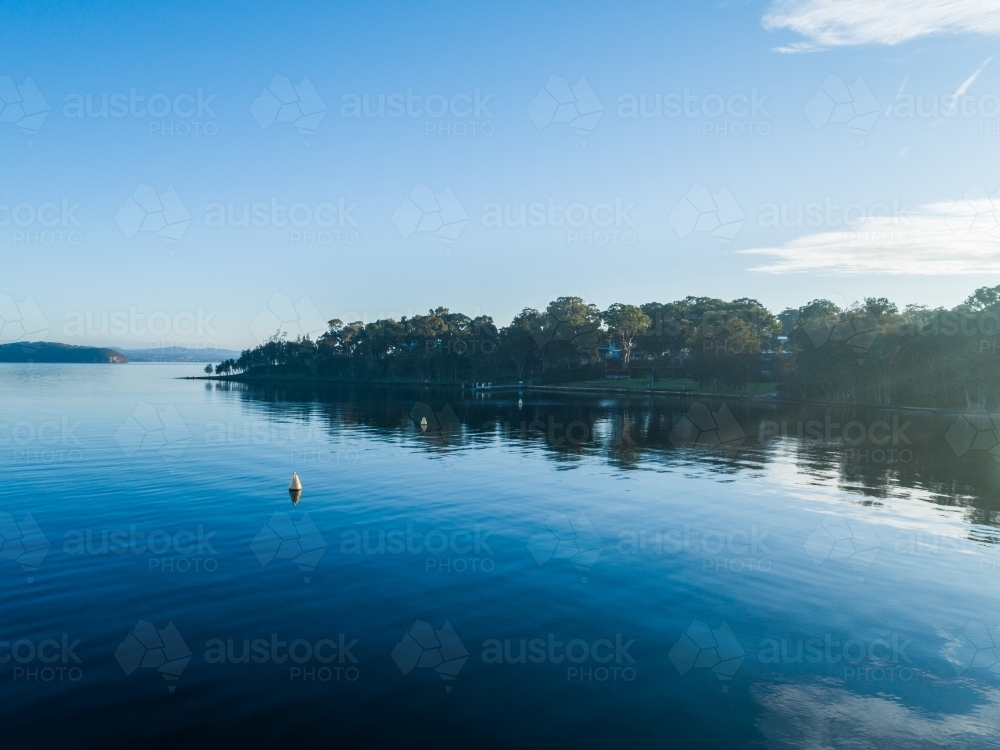 Trees on shoreline of lake with water reflecting blue sky on cold morning seen from aerial view - Australian Stock Image