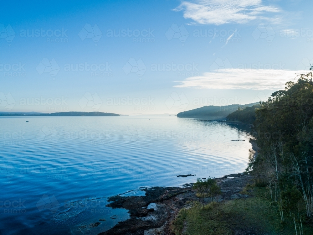 Trees on shoreline of lake with water reflecting blue sky on cold morning seen from aerial view - Australian Stock Image