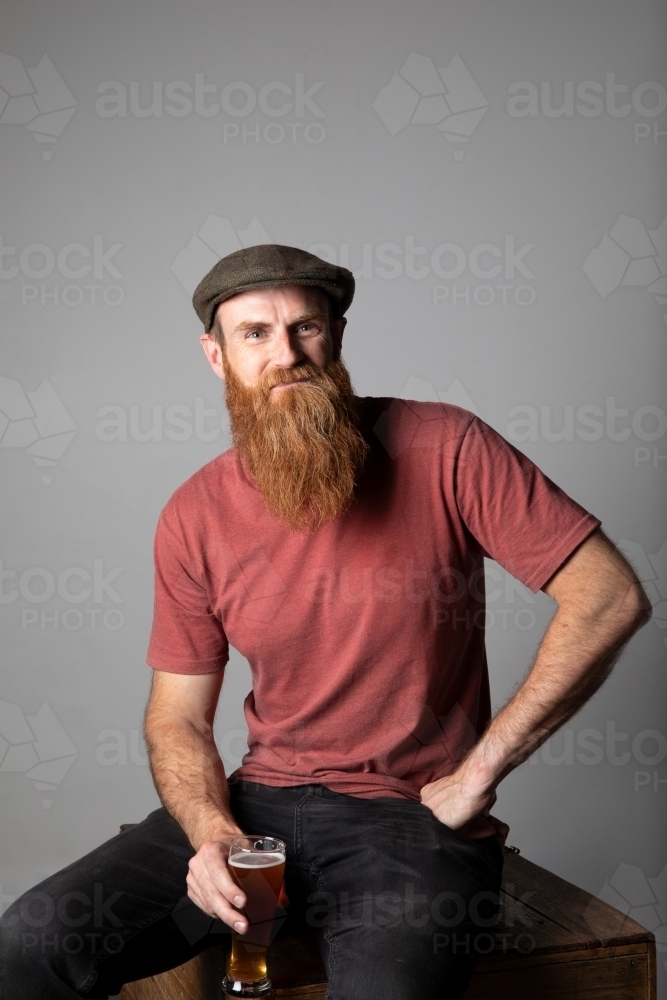 Trendy man with ginger beard and flat cap, holding a glass of beer - Australian Stock Image