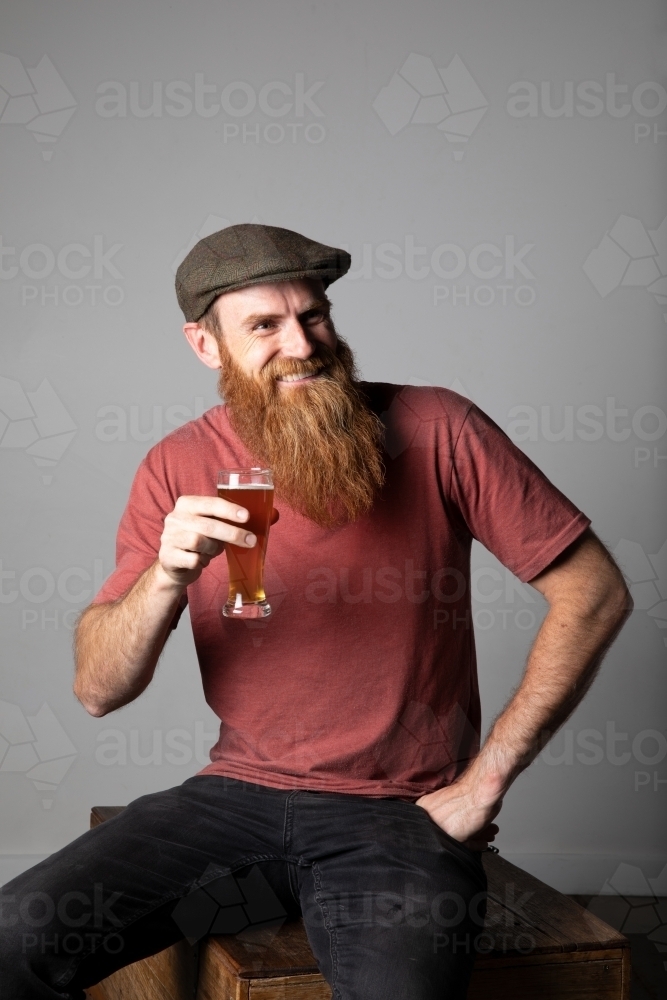 Trendy man with ginger beard and flat cap, holding a glass of beer - Australian Stock Image