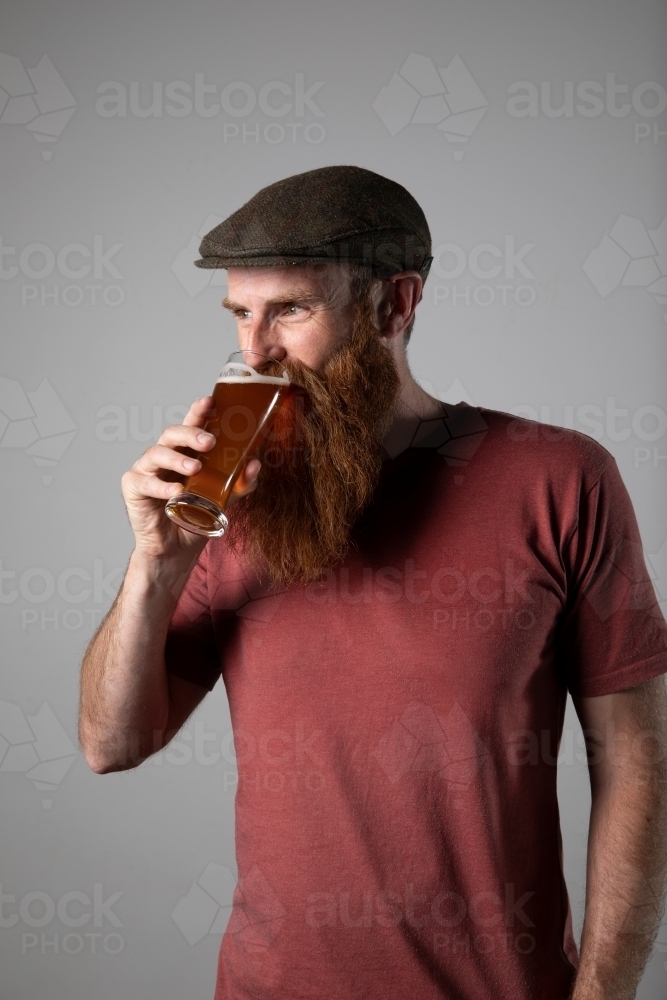 Trendy man with ginger beard and flat cap, holding a glass of beer - Australian Stock Image