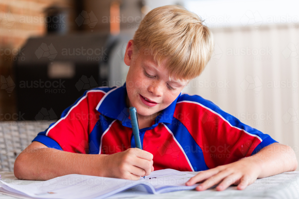 Tween boy in school uniform doing bookwork homework on table in backyard at home - Australian Stock Image