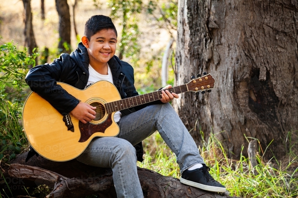 Tween boy playing guitar in bushland seated on log - Australian Stock Image
