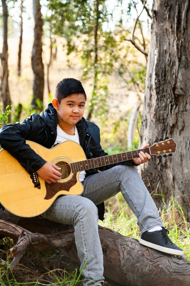 Tween boy playing guitar in bushland seated on log - Australian Stock Image