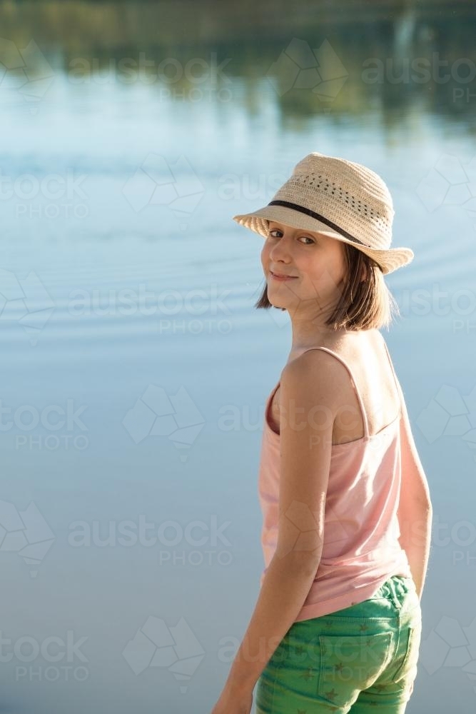 Tween girl by a river in late afternoon sunlight, smiling back to camera - Australian Stock Image