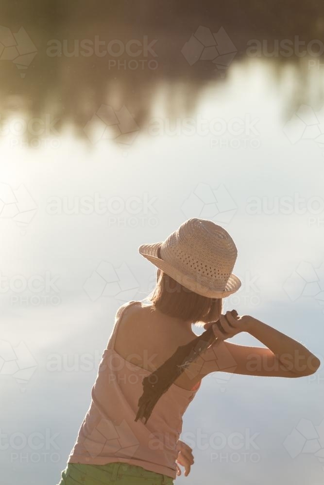 Tween girl by a river in late afternoon sunlight, throwing a stick into the water - Australian Stock Image