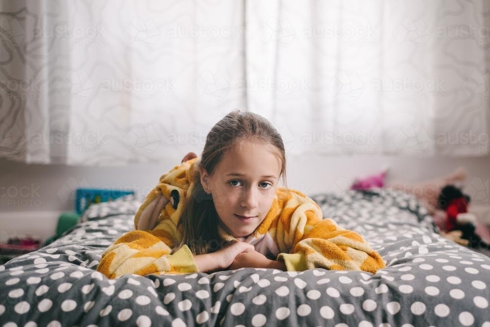 Tween girl in a giraffe onesie lying on her bed, looking to camera - Australian Stock Image
