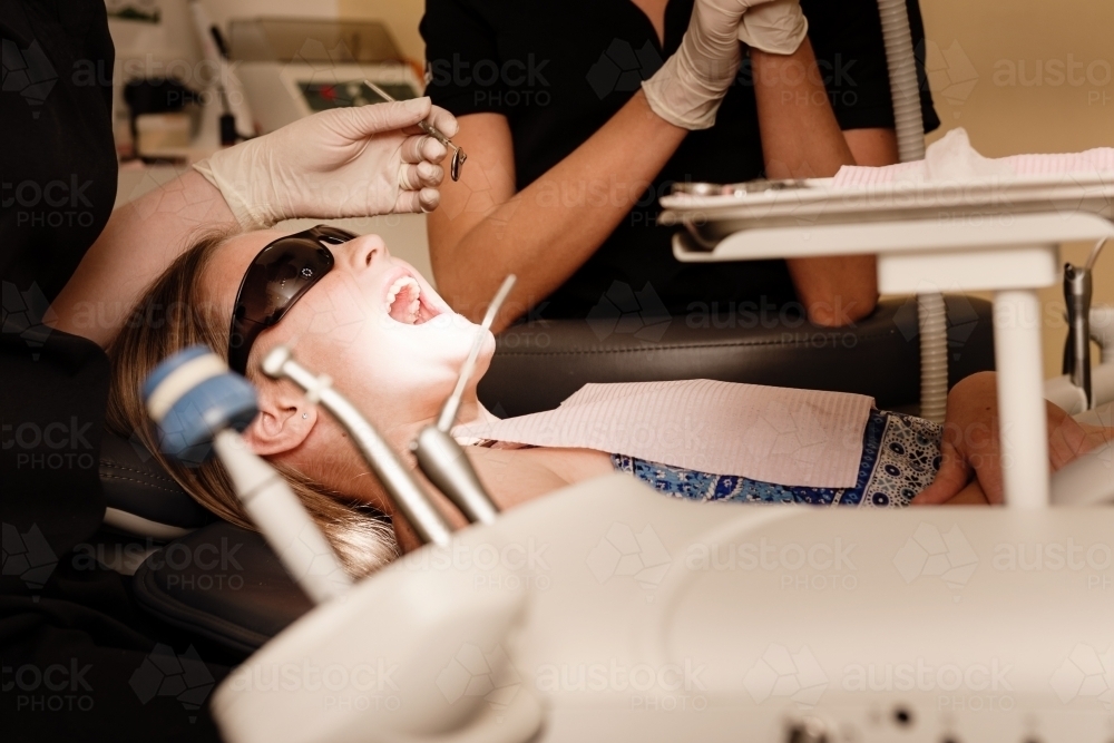 Tween girl lying in the dentist chair, with bright light on her mouth - Australian Stock Image