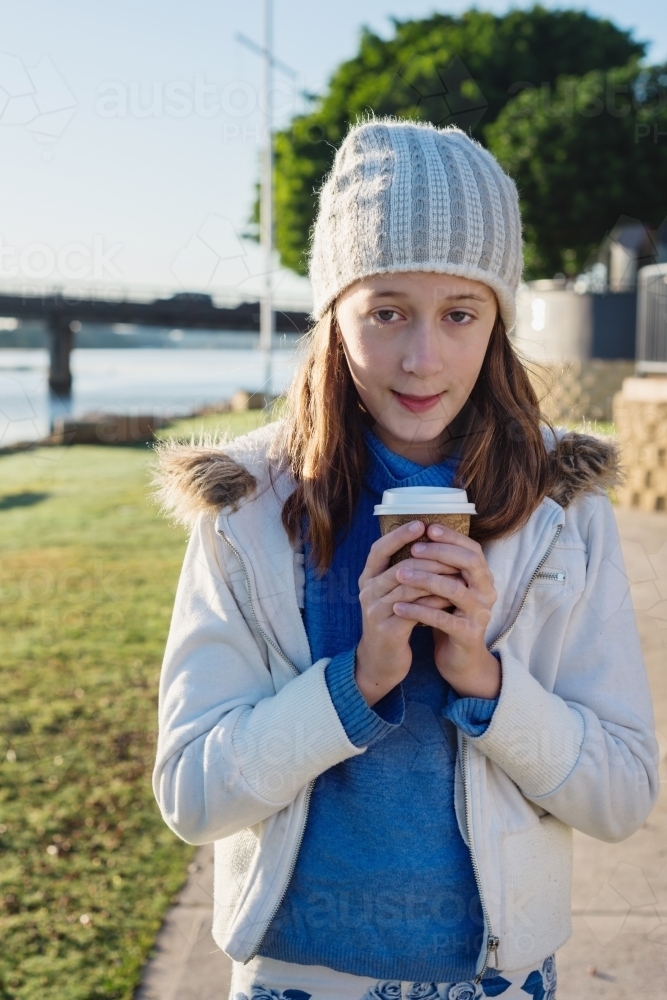 tween girl wearing beanie in winter, by river with a hot chocolate - Australian Stock Image