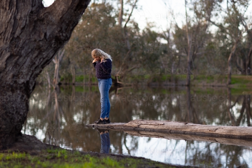 tween girl wearing jeans looking at reflection in water standing on log - Australian Stock Image