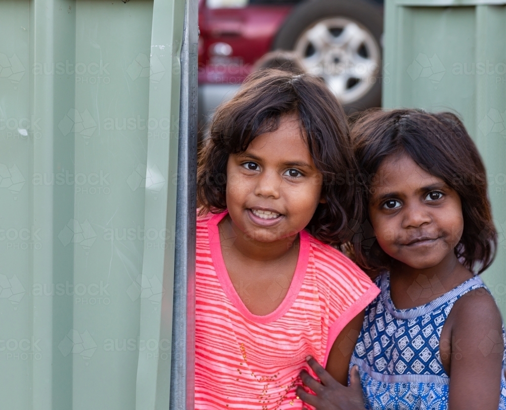 Two Aboriginal children looking out of open gate in tin fence - Australian Stock Image