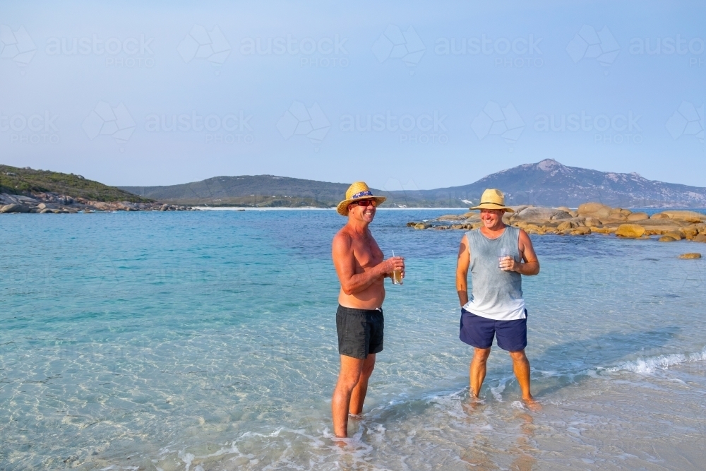 two blokes standing in the water holding drinks at the beach - Australian Stock Image