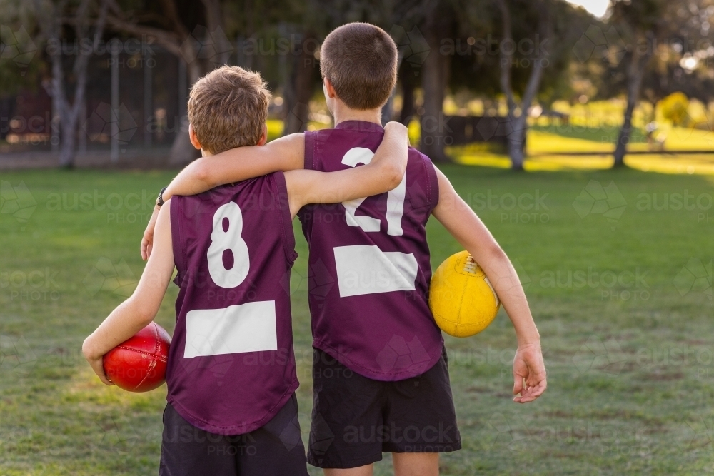 two boys in football gear seen from behind holding footballs - Australian Stock Image