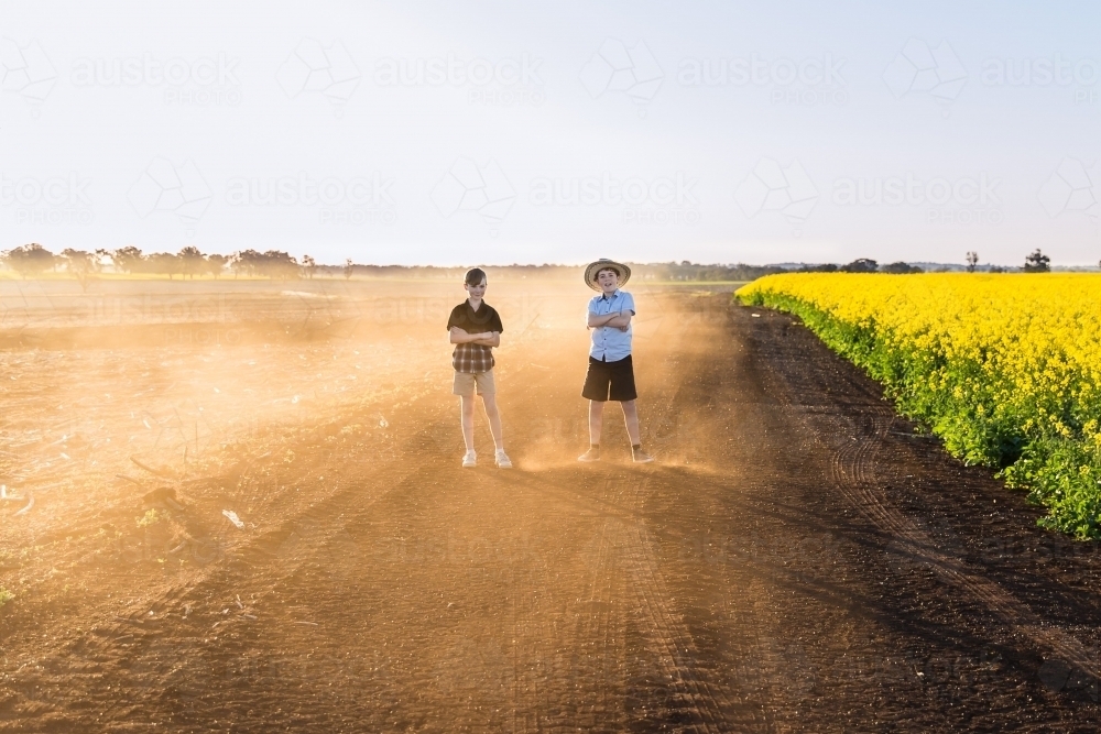 Two boys standing with arms crossed on dusty dirt road next to canola field on farm - Australian Stock Image