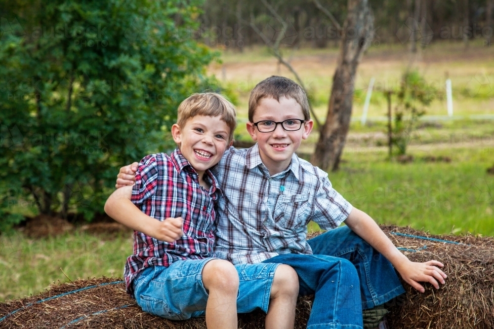 Two brothers sitting together on hay bales grinning - Australian Stock Image