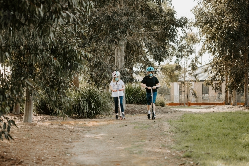 two Caucasian children riding scooters wearing masks during the Corona COVID-19 pandemic - Australian Stock Image