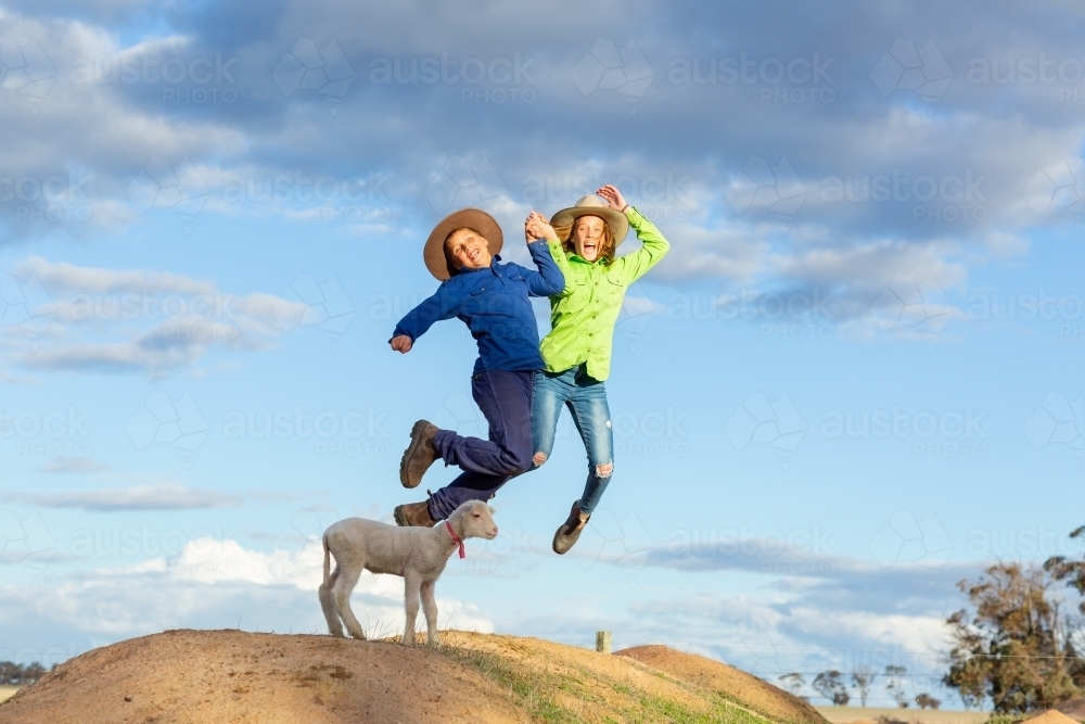 Two children jumping with pet lamb on mound - Australian Stock Image