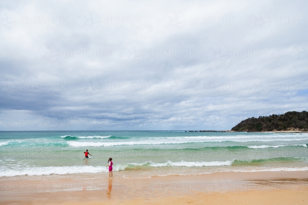 Two distant children playing in the beach waves  on overcast day - Australian Stock Image