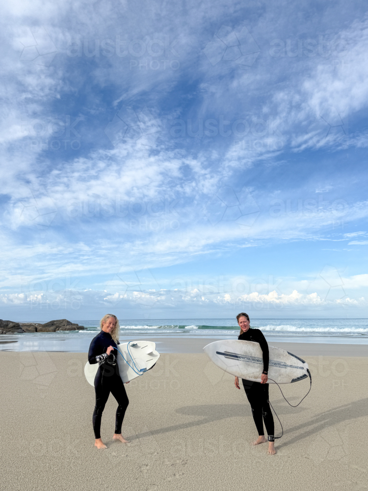 Two female surfers standing on beach side by side looking out to ocean in wetsuits - Australian Stock Image