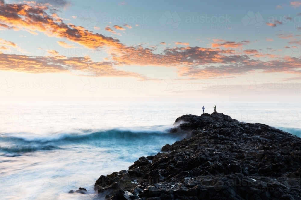 Two fishermen fish in the Pacific Ocean from a rocky headland in a long exposure sunrise scene - Australian Stock Image