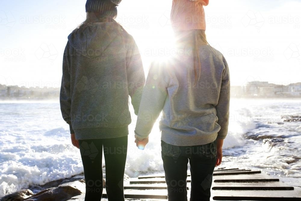 Two girls hold hands on a boat ramp by the ocean in the afternoon light - Australian Stock Image