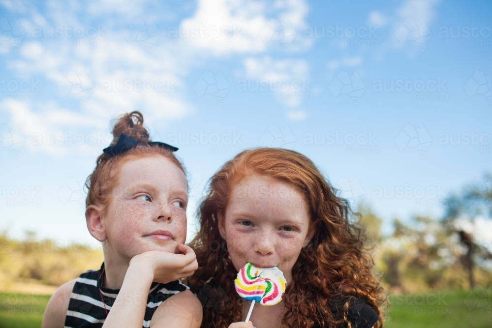 Two girls outside eating a rainbow lollipop - Australian Stock Image
