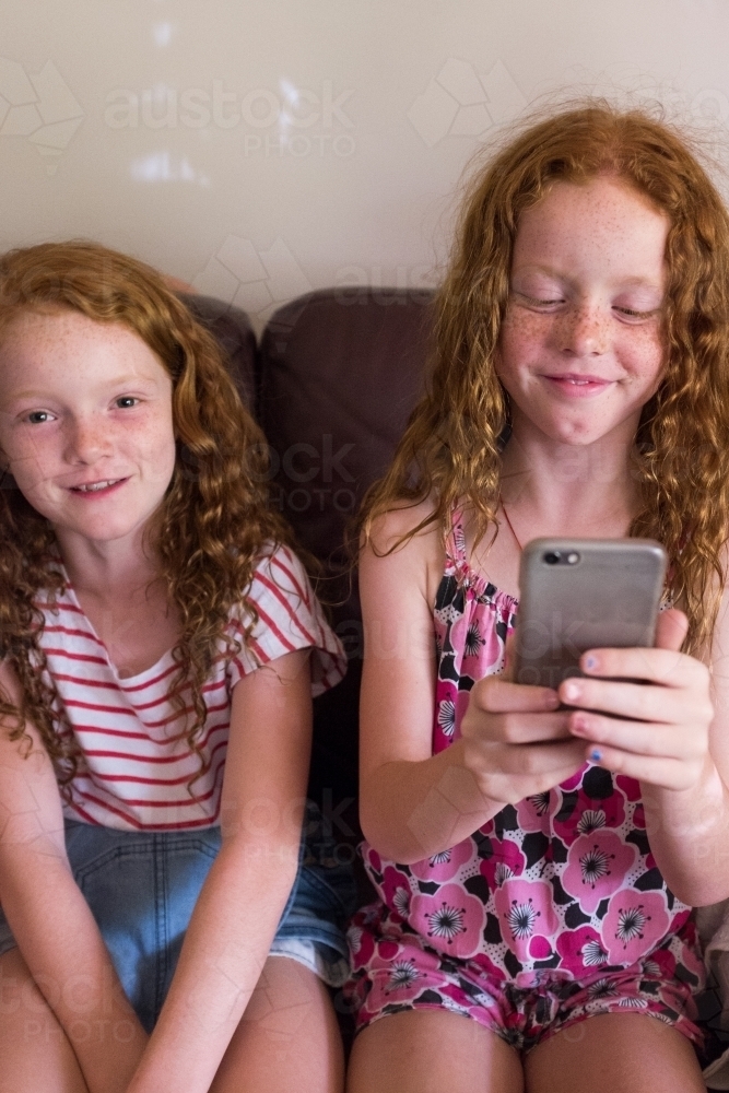 Two girls sitting on a couch playing on a smartphone - Australian Stock Image